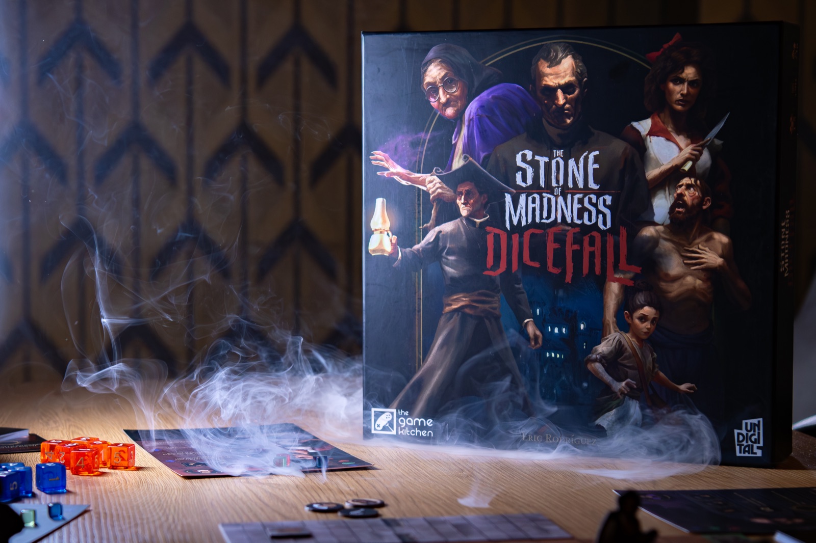 Official Gamefound photo of The Stone of Madness: Dicefall box beside orange and blue dice on a wooden table.