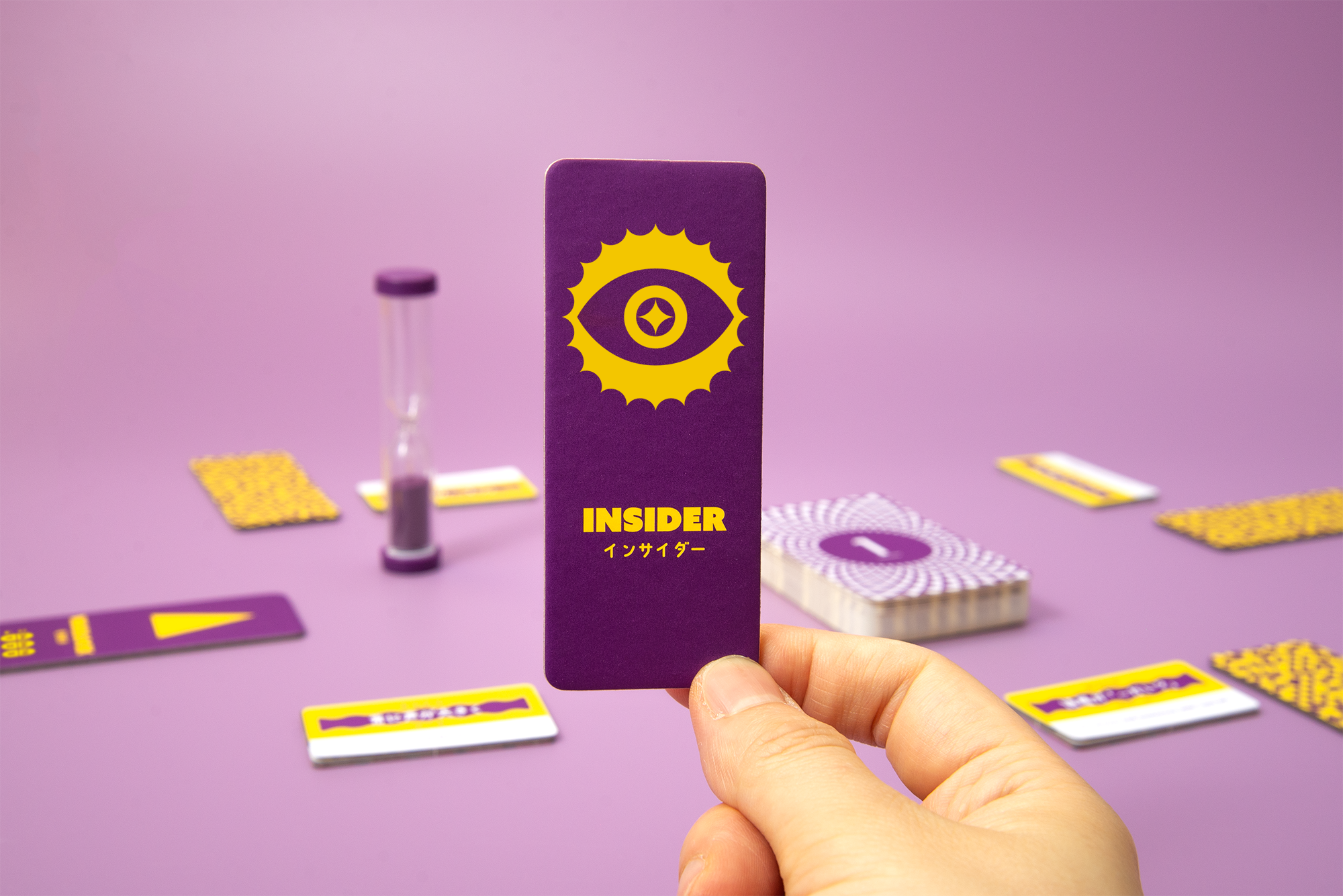 A hand holds the purple Insider role card beside the sand timer and magic cards on a pink table.